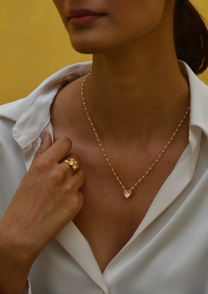 Close-up of a woman displaying a heart-shaped pendant and elegant ring against a yellow backdrop.